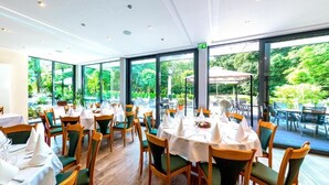 Ein Restaurant mit runden Tischen und Stühlen, großen Fenstern mit Blick auf Grünflächen und einer Decke mit eingelassener Beleuchtung.