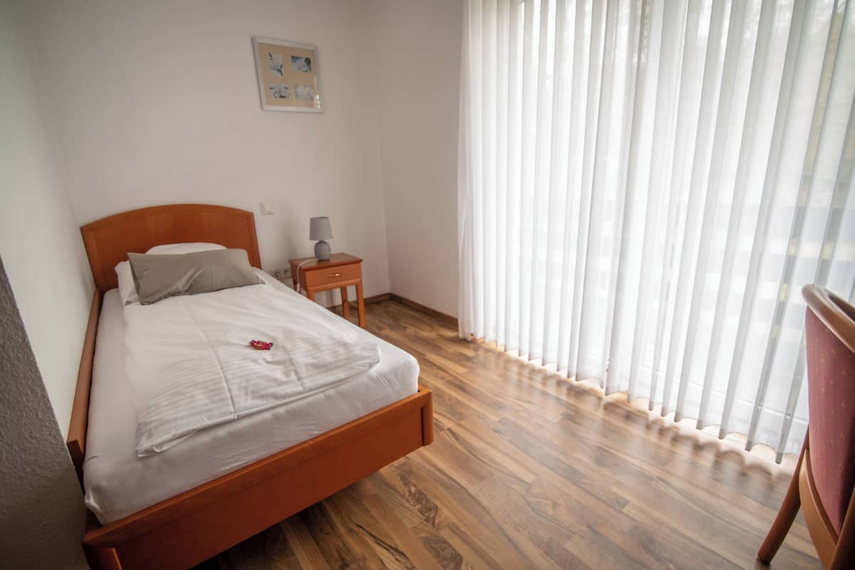 Una cama bien tendida con cabecera de madera, una mesita de noche con una lámpara y una ventana con cortinas translúcidas.