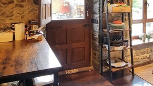 Un interior rústico con pared de piedra, vigas de madera en el techo, una mesa de madera y un estante de piedra con utensilios de cocina.