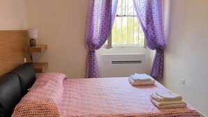 A bedroom with a bed, a bedside table with a lamp, a window with purple curtains, and an air conditioner. - Casa Mariuccia Cozy apartment near the waterfront Valencia (Alghero)