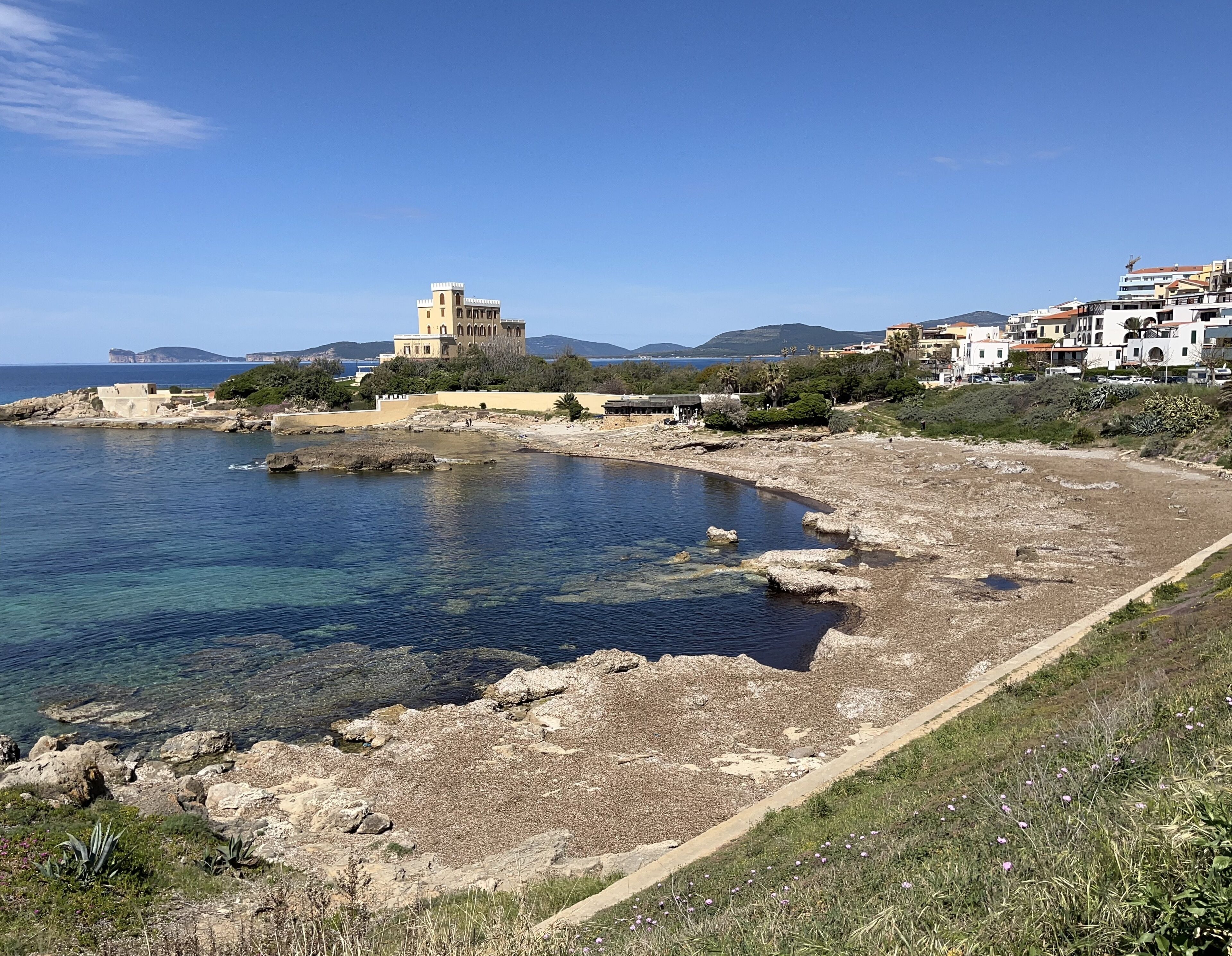 A coastal area with a clear view of the sea, rocky beach, and a historic building with a tower.