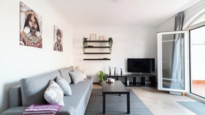 Ett modernt vardagsrum med en grå soffa, ett svart soffbord och en tv som står på ett tv-bord. Det finns hyllor med växter och tavlor på väggen.
