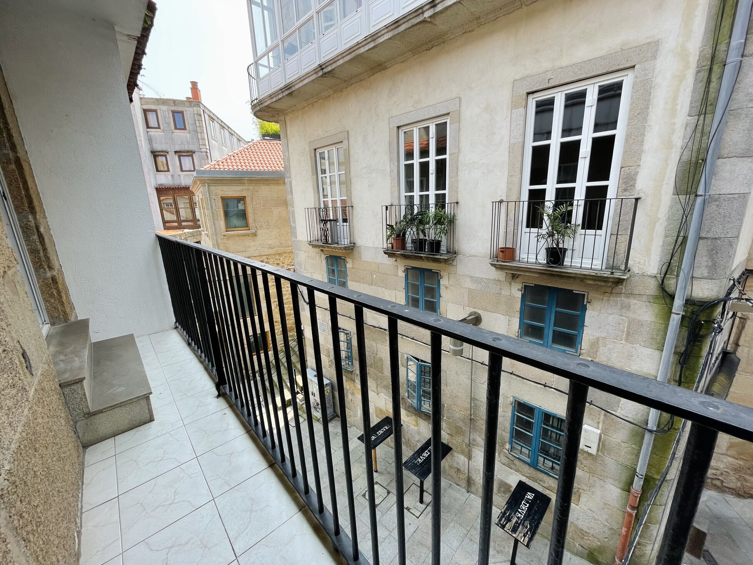 Een balkon met betegelde vloer en een zwarte metalen reling. Op de vensterbanken van het aangrenzende gebouw staan potplanten.