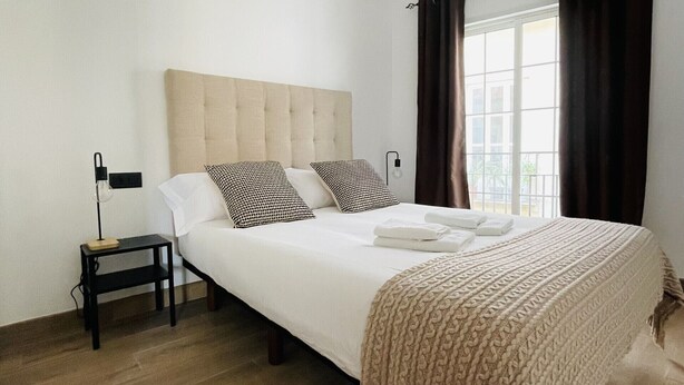 Een slaapkamer met een bed, nachtkastje, lamp en raam met gordijnen.
