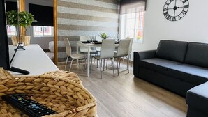 Een moderne woonkamer met een bank, een eettafel met stoelen, een gevlochten mand met een afstandsbediening, en een wandklok.