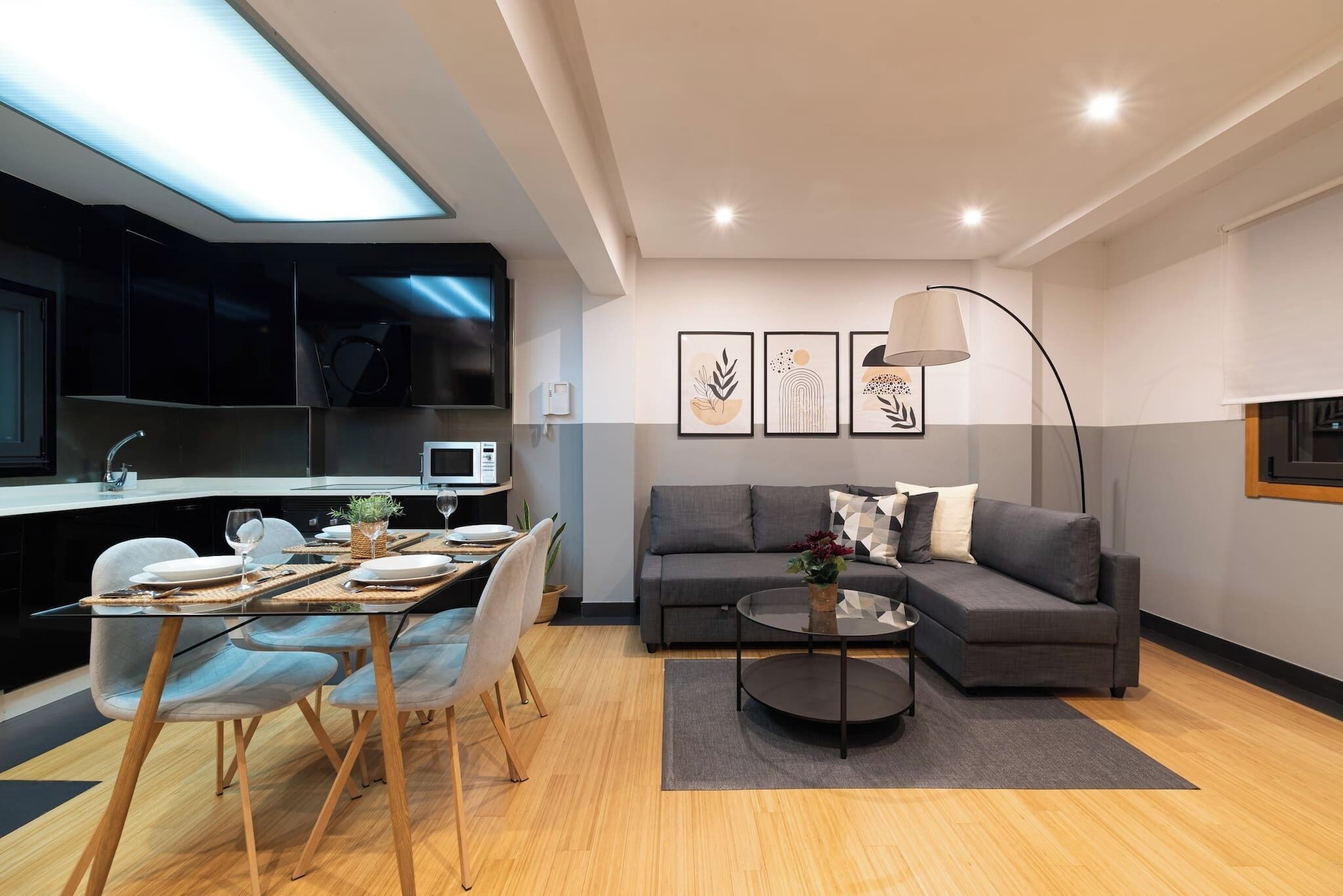 A modern living room with a grey sofa, a glass dining table, and a kitchen area with a TV and microwave.