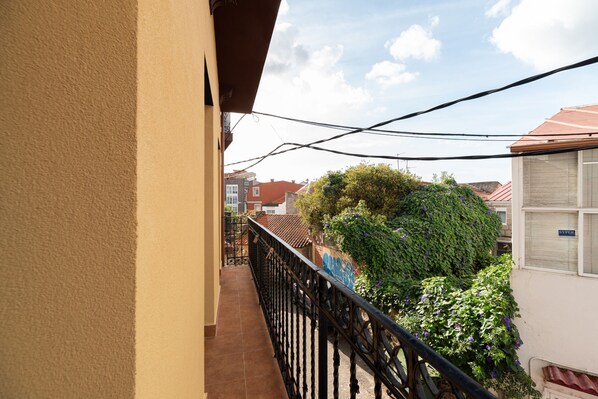 Un balcon doté d’une balustrade en métal noir, d’un sol carrelé et offrant une vue sur les bâtiments voisins et la verdure environnante.