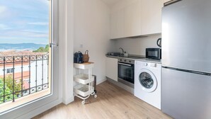 A compact kitchen with a washing machine, oven, and microwave. A small cart with baskets is near the window, which offers a view of buildings and greenery.
