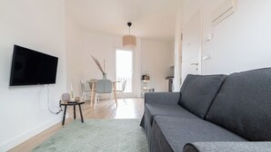 A modern living room with a grey sofa, a flat-screen TV mounted on the wall, a small side table with a vase, and a patterned rug.