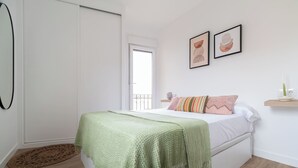 A bedroom with a bed, a green blanket, a wooden floor, a white door, and a window.