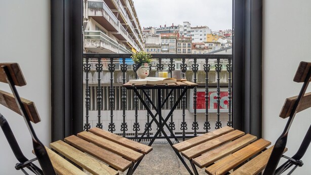 Un balcon avec deux chaises en bois, une petite table sur laquelle se trouvent un livre et un vase de fleurs, et une vue sur un paysage urbain avec des bâtiments.
