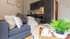 Un salon moderne avec un canapé gris, une table basse agrémentée d’une plante, et une cuisine en arrière-plan.