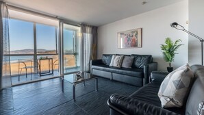 En moderne stue med en sort lædersofa, et glasbord og udsigt til stranden gennem store vinduer.