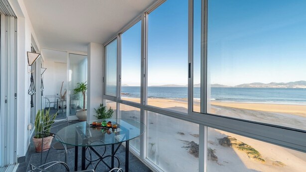 Et moderne værelse med et glasbord og stole, store vinduer med udsigt til en strand.