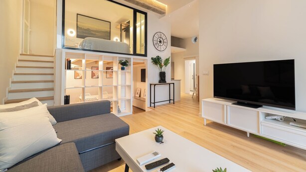 Una sala de estar moderna con un sofá gris, una mesa de centro blanca y un televisor de pantalla plana grande sobre un soporte blanco. Se observa una escalera de madera que lleva a un nivel superior, y una pared blanca con un reloj y un cuadro enmarcado.