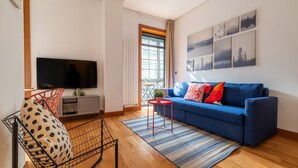 A living room with a blue sofa, a flat-screen TV, a red chair, and a window with a view of trees. - La Galeria de Principe (Vigo)