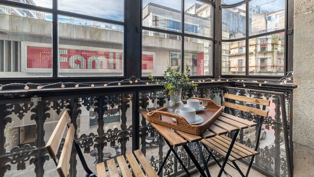 A balcony with a wooden table, wicker chairs, and a vase with flowers. - La Galeria de Principe (Vigo)