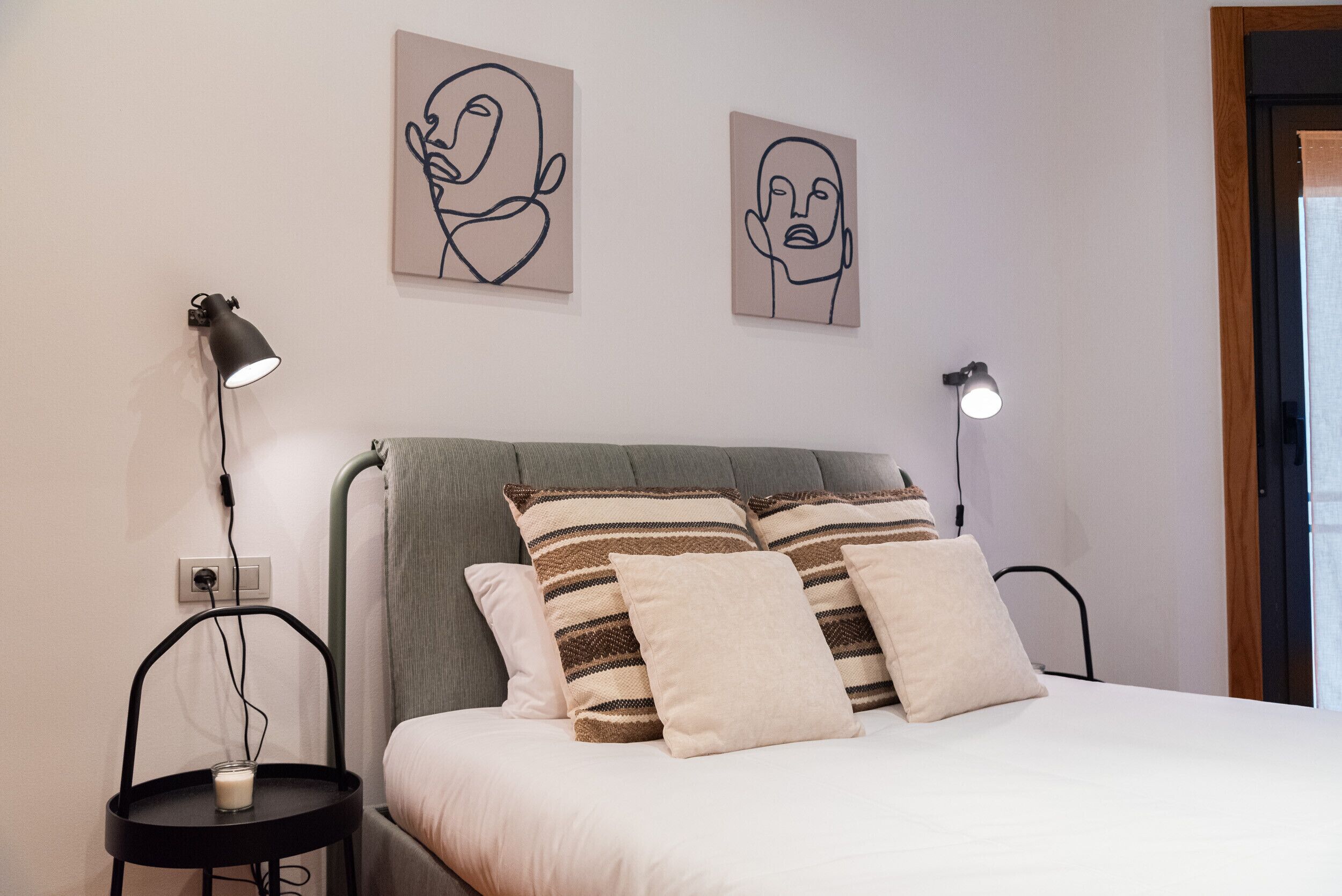 En pænt redt seng med stribede puder, en sengetavlelampe og en lampe på væggen.