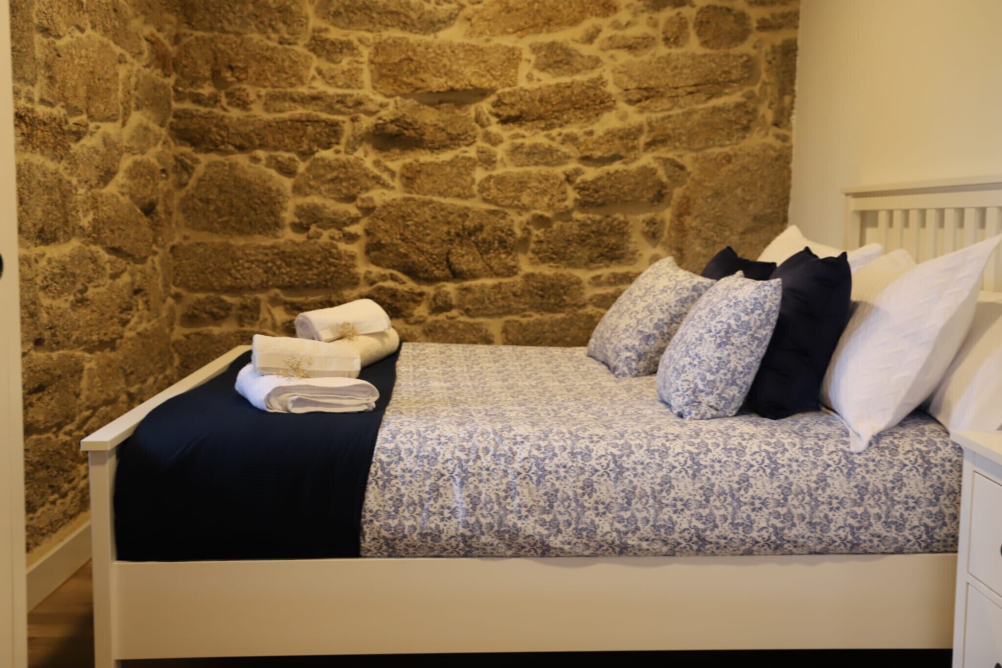 A bed with a floral bedspread, white pillows, and a rolled towel on a white frame against a stone wall.