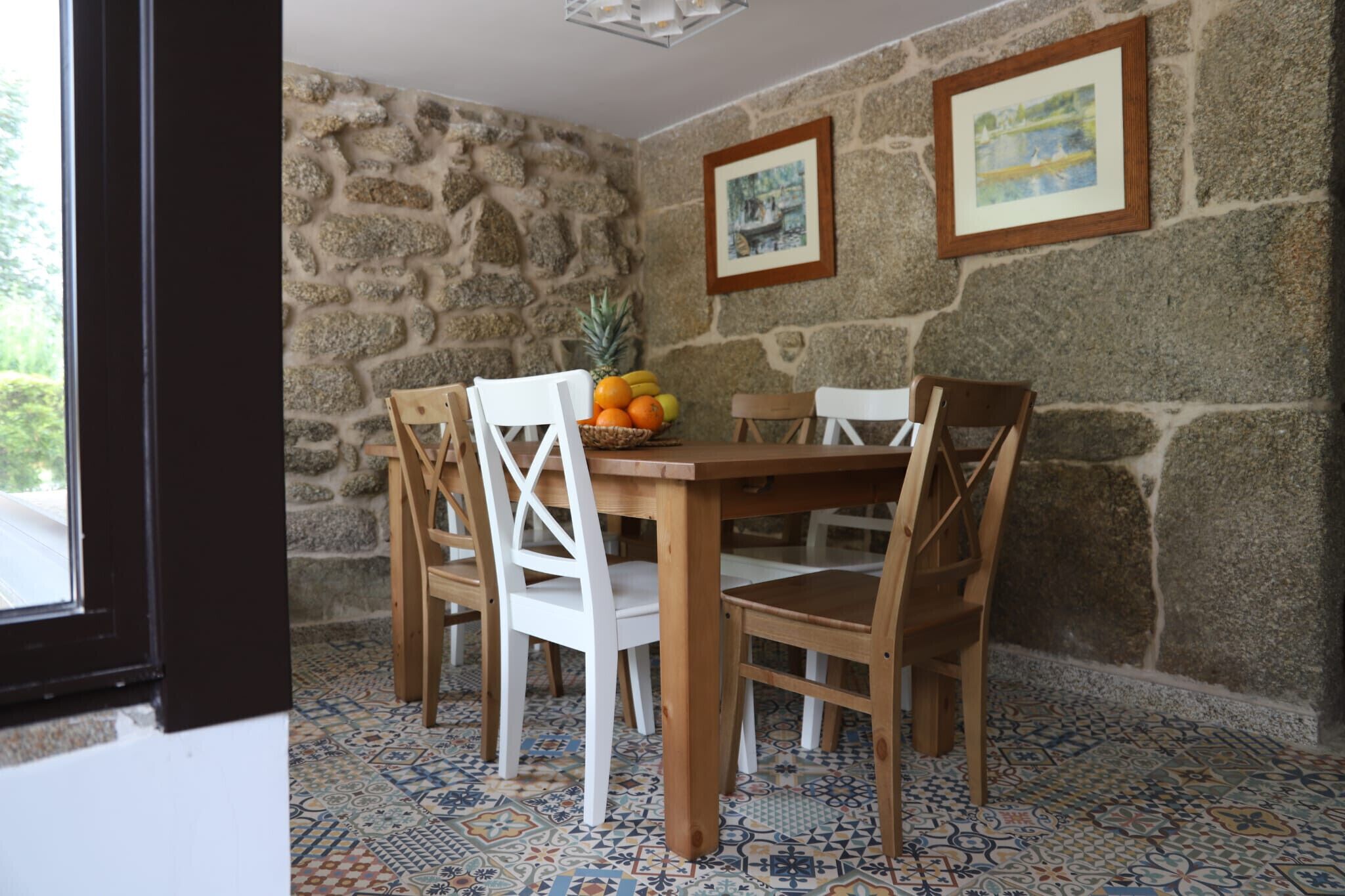 A dining area with a stone wall, wooden table, white chairs, and a fruit basket.