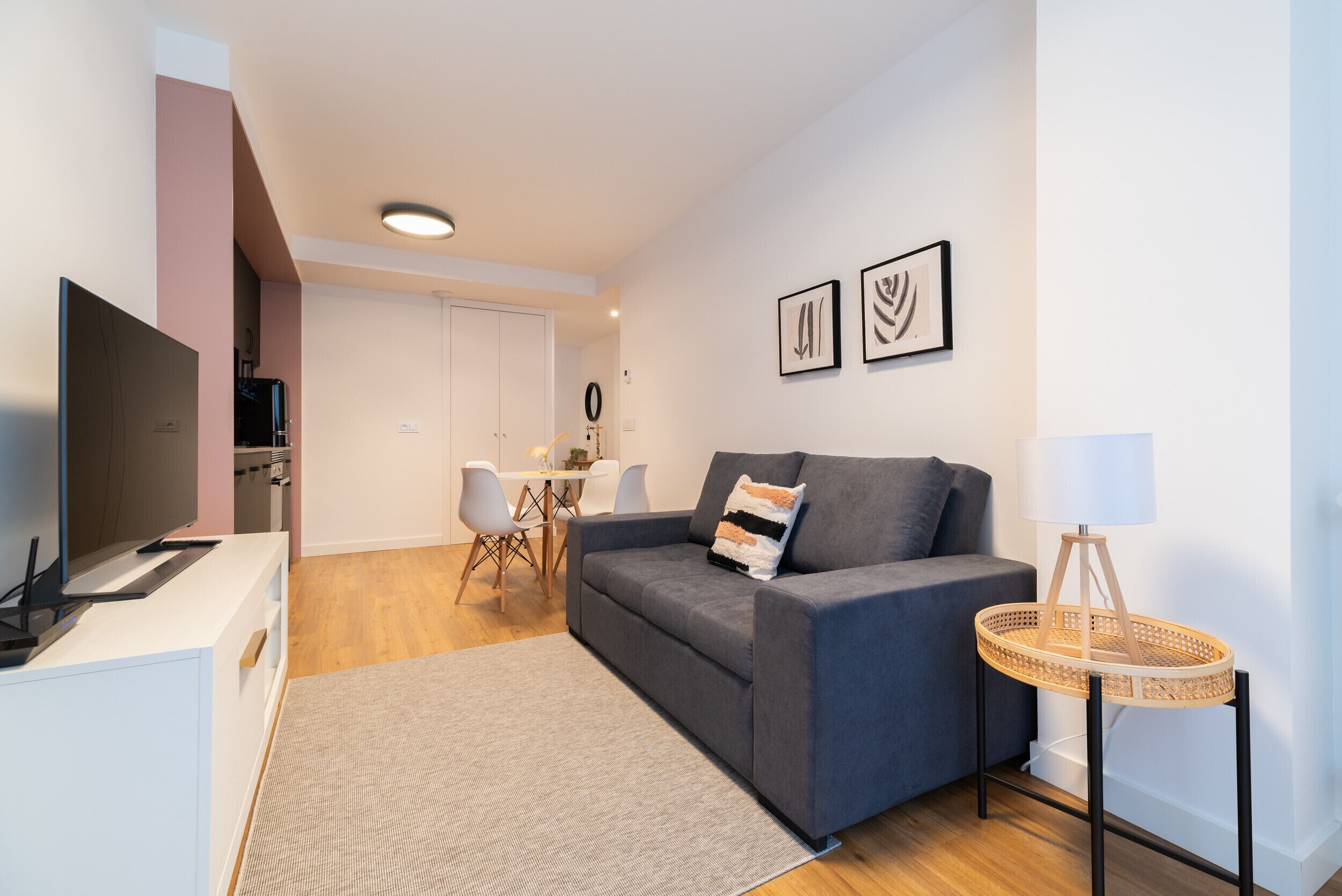 A modern living room with a grey sofa, a TV, a small dining table, and a lamp.