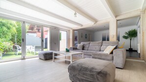 A spacious living area with a grey sofa, a white coffee table, and a large grey ottoman. There is a sliding glass door leading to an outdoor patio with a table and chairs. - House Vigo (Vigo)