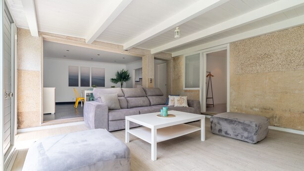A modern living room with a sofa, ottoman, and coffee table. There is a door leading to another room, and a plant is visible in the background. - House Vigo (Vigo)