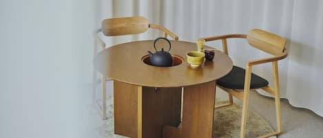 Ein runder Holztisch mit einem schwarzen Wasserkocher, einer gelben Tasse und einer kleinen, dunklen Tasse. Zwei Holzsessel mit schwarzen Kissen befinden sich um den Tisch herum.