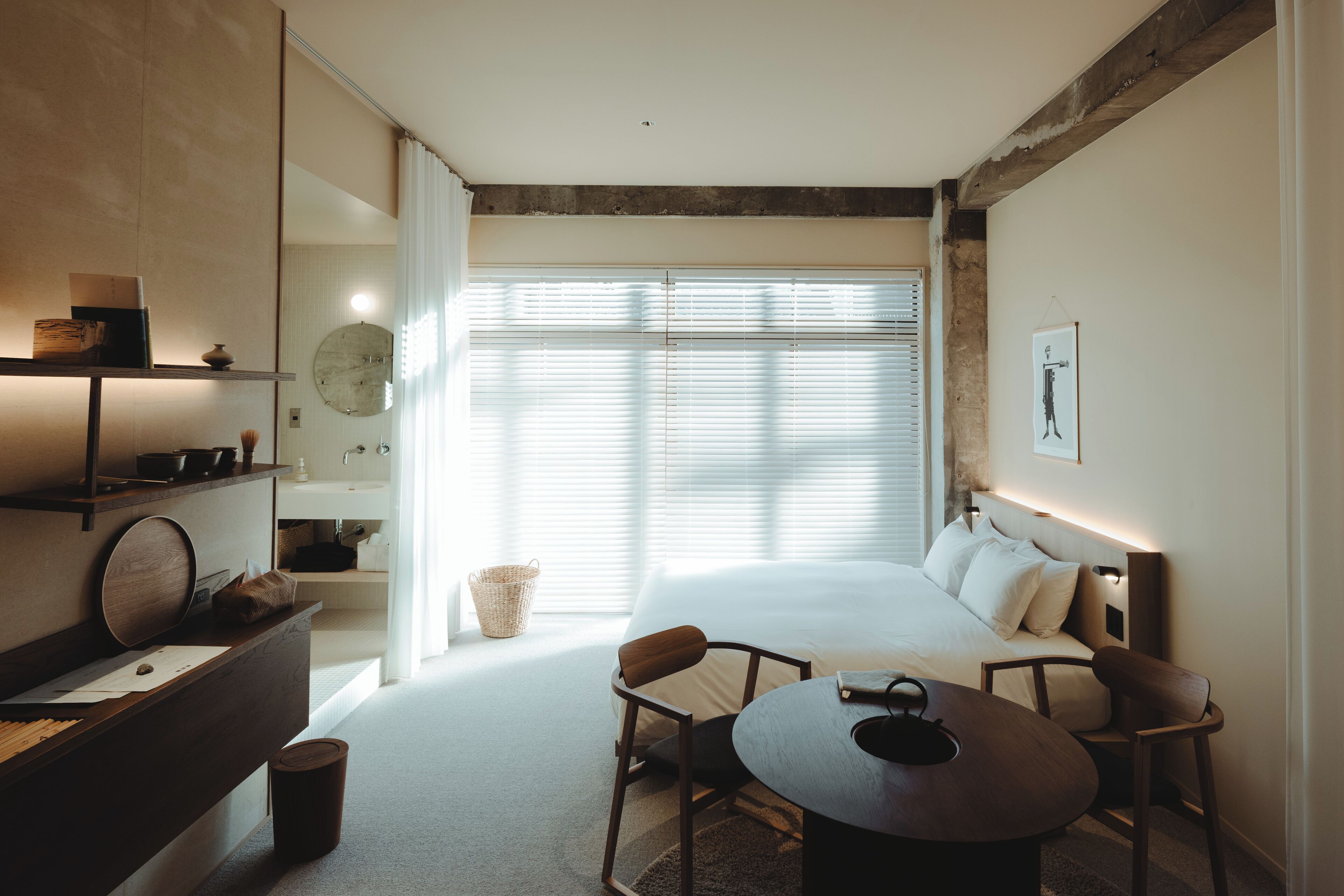 A modern hotel room with a large bed, a desk with a chair, a small table, and a window with blinds.