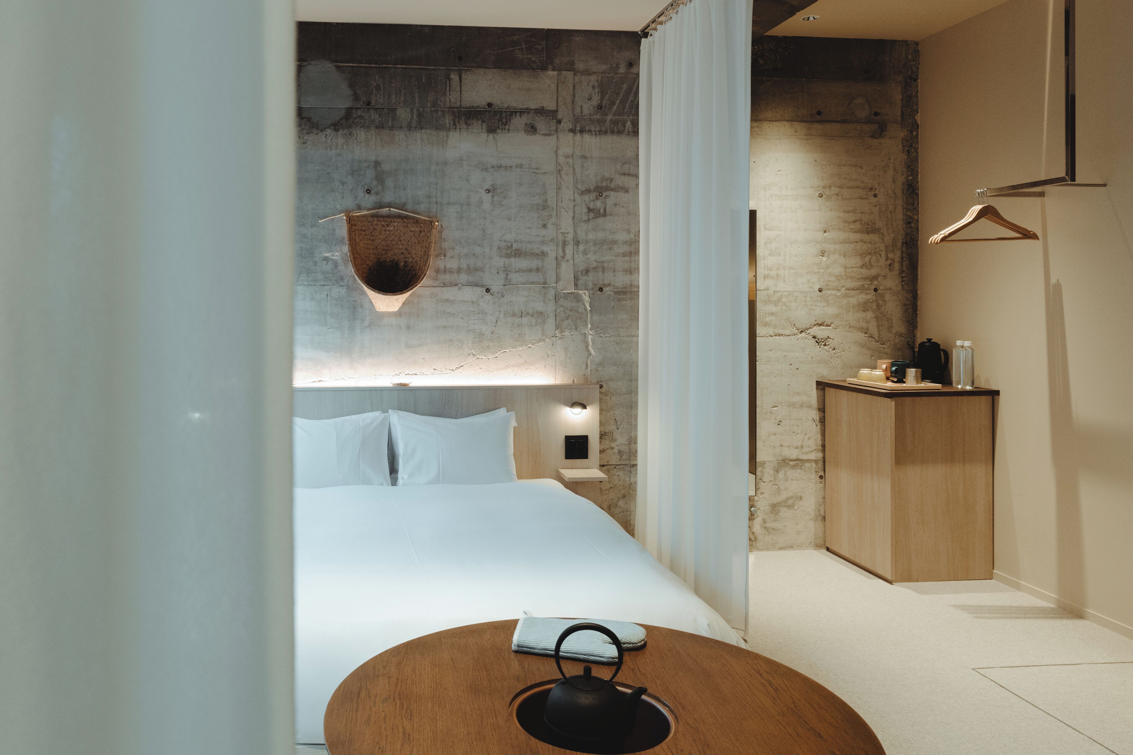 Quarto de hotel moderno com uma cama grande, uma mesa de cabeceira de madeira e uma parede de concreto como detalhe.