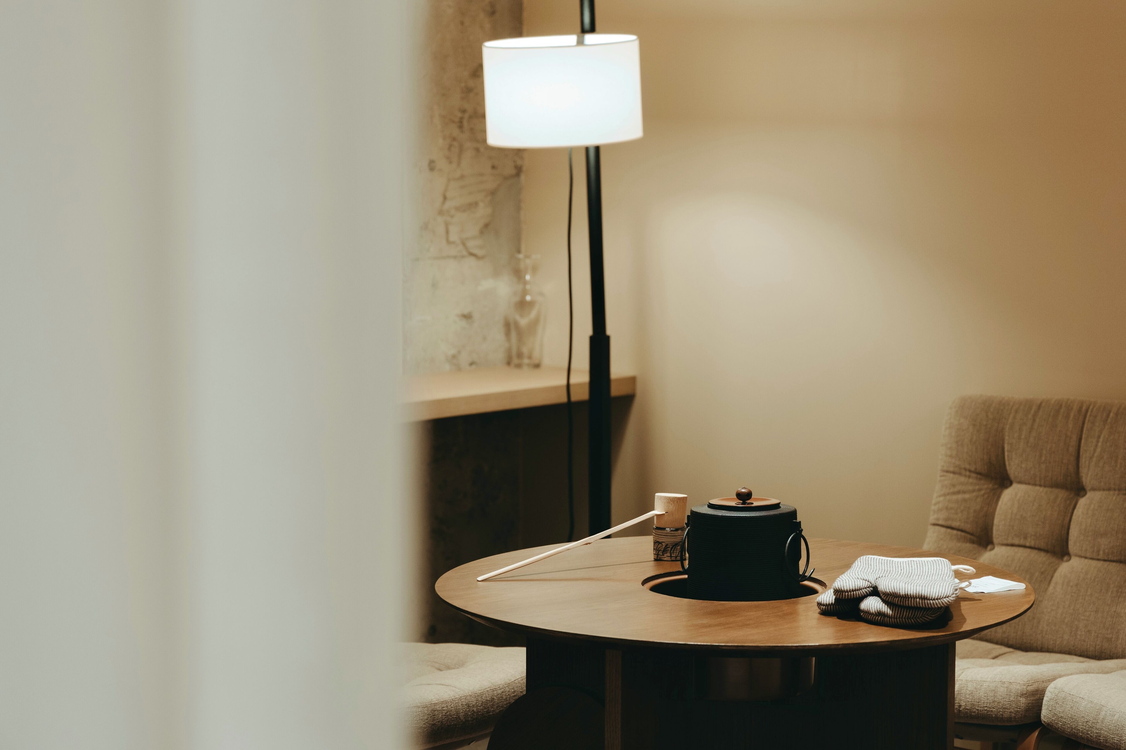 A round wooden table with a lamp, a small bottle, a cup, and a folded towel. There is a sofa and a wall with a textured pattern in the background.