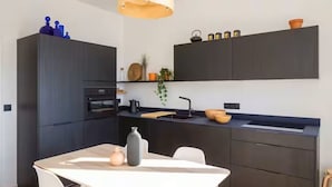 A modern kitchen with dark cabinetry, a built-in oven, and a sink. There is a dining table with chairs and a vase on it. - APARTMENT WITH MAGNIFICENT VIEW OF THE OLD PORT (Marseille)
