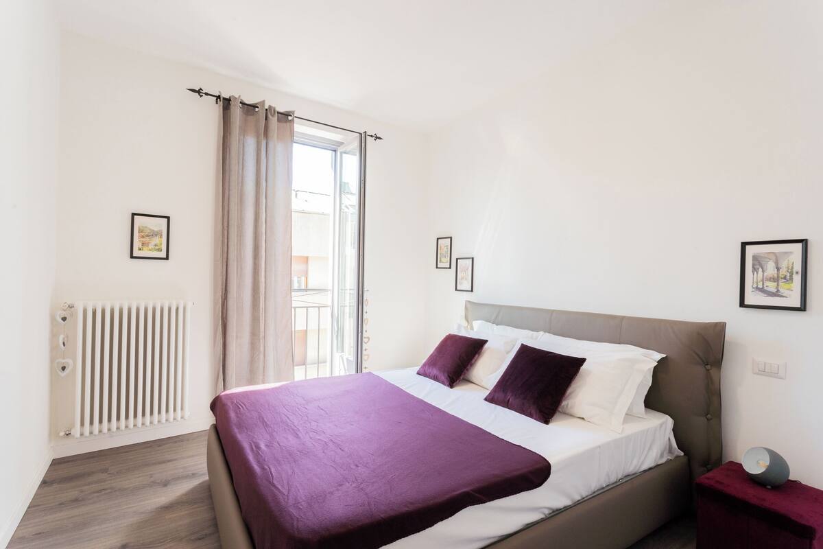 Ein Schlafzimmer mit einem Bett, einem Heizkörper, einem Fenster mit Vorhängen und einem Nachttisch mit Lampe.