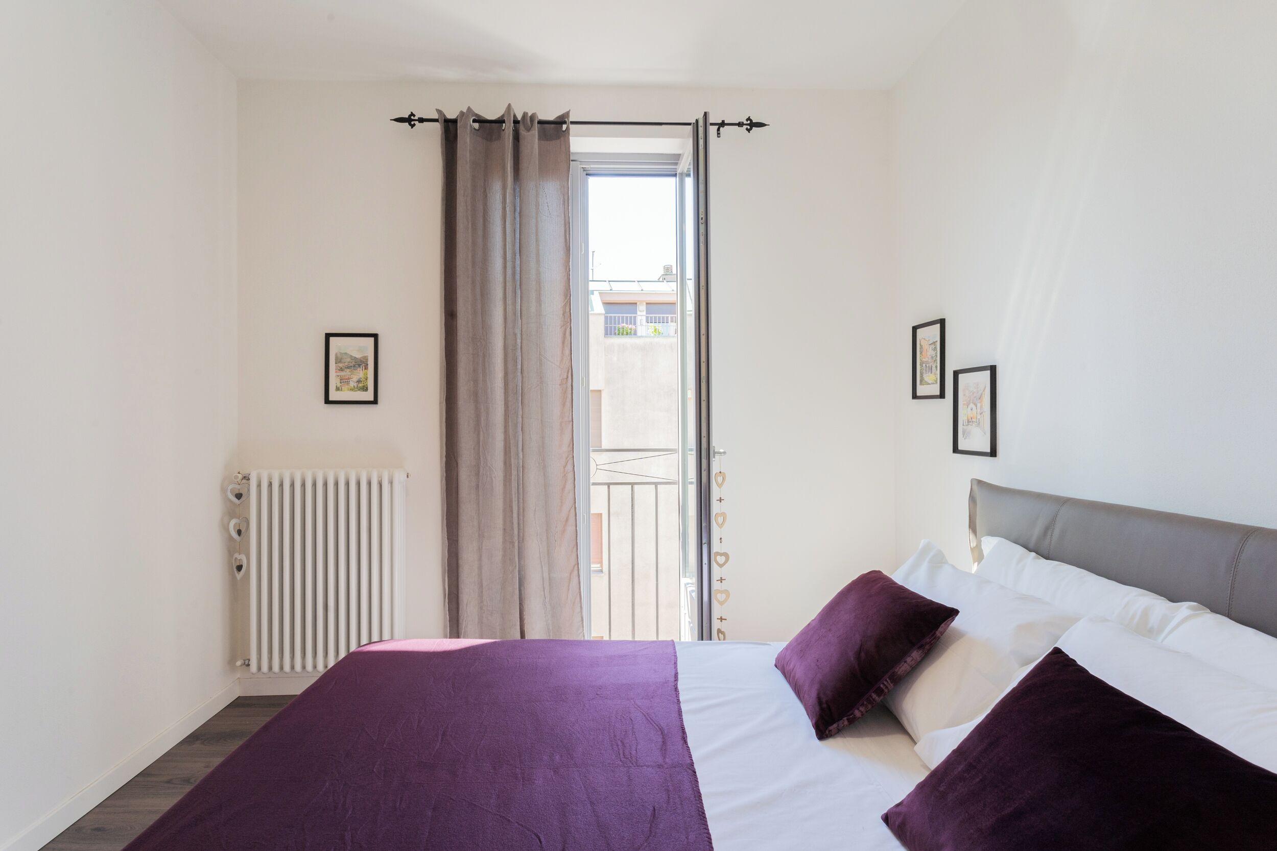 A bedroom with a bed, a radiator, a window with curtains, and a balcony view.