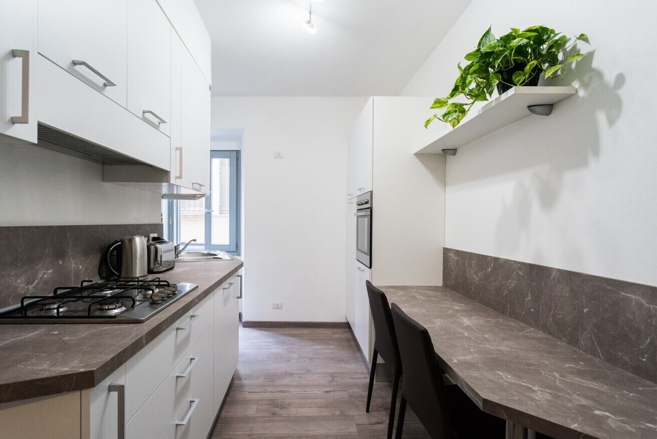 A modern kitchen with white cabinets, a gas stove, and a marble countertop. There is a plant on a shelf above the counter and a window providing natural light.