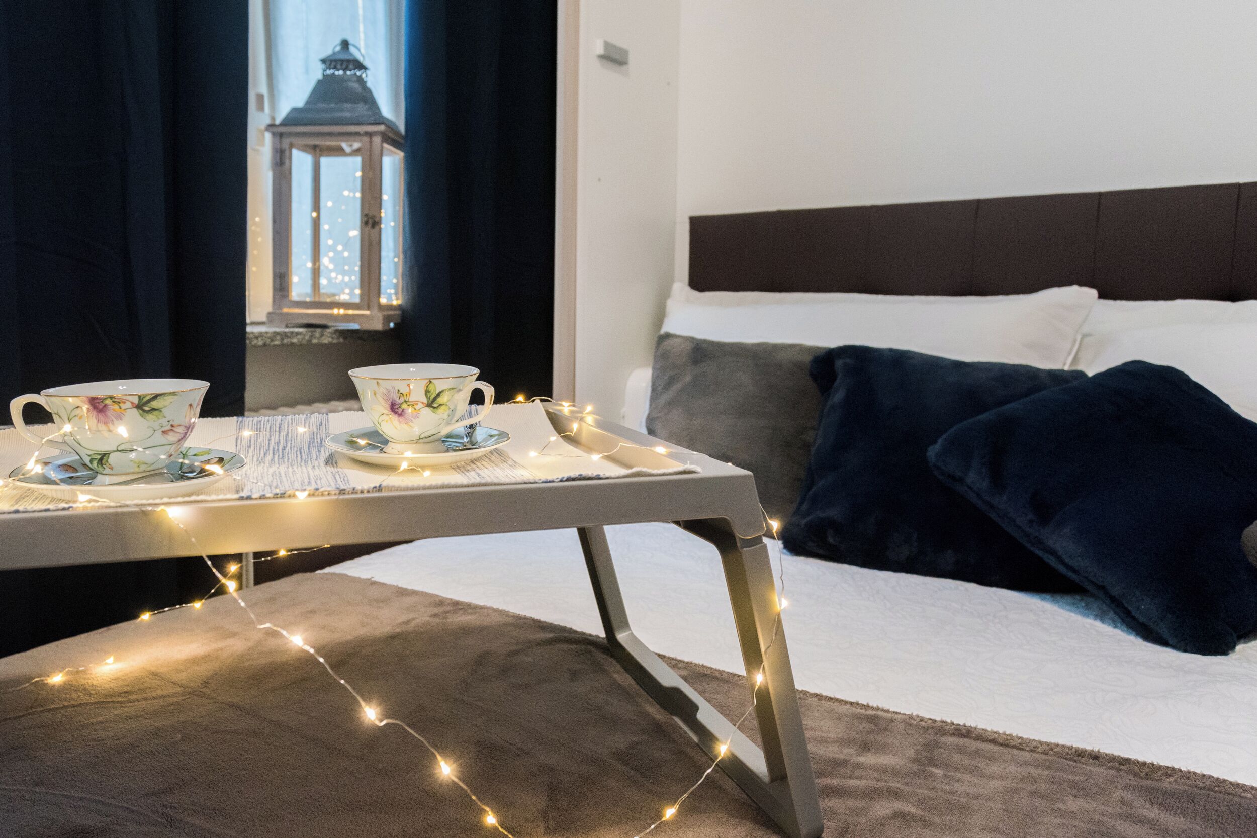 Un lit bien fait, avec une tête de lit sombre, une table de chevet sur laquelle se trouve un plateau avec des tasses et des soucoupes à thé, et une guirlande de petites lumières scintillantes.