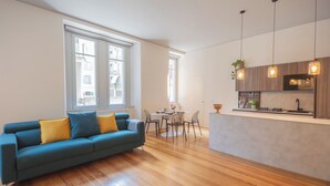 A modern living room with a blue sofa, wooden flooring, a dining area with a table and chairs, and a kitchen with pendant lights and a microwave. - Como Liberty Suites - Garbo by Rent all Como (Como)