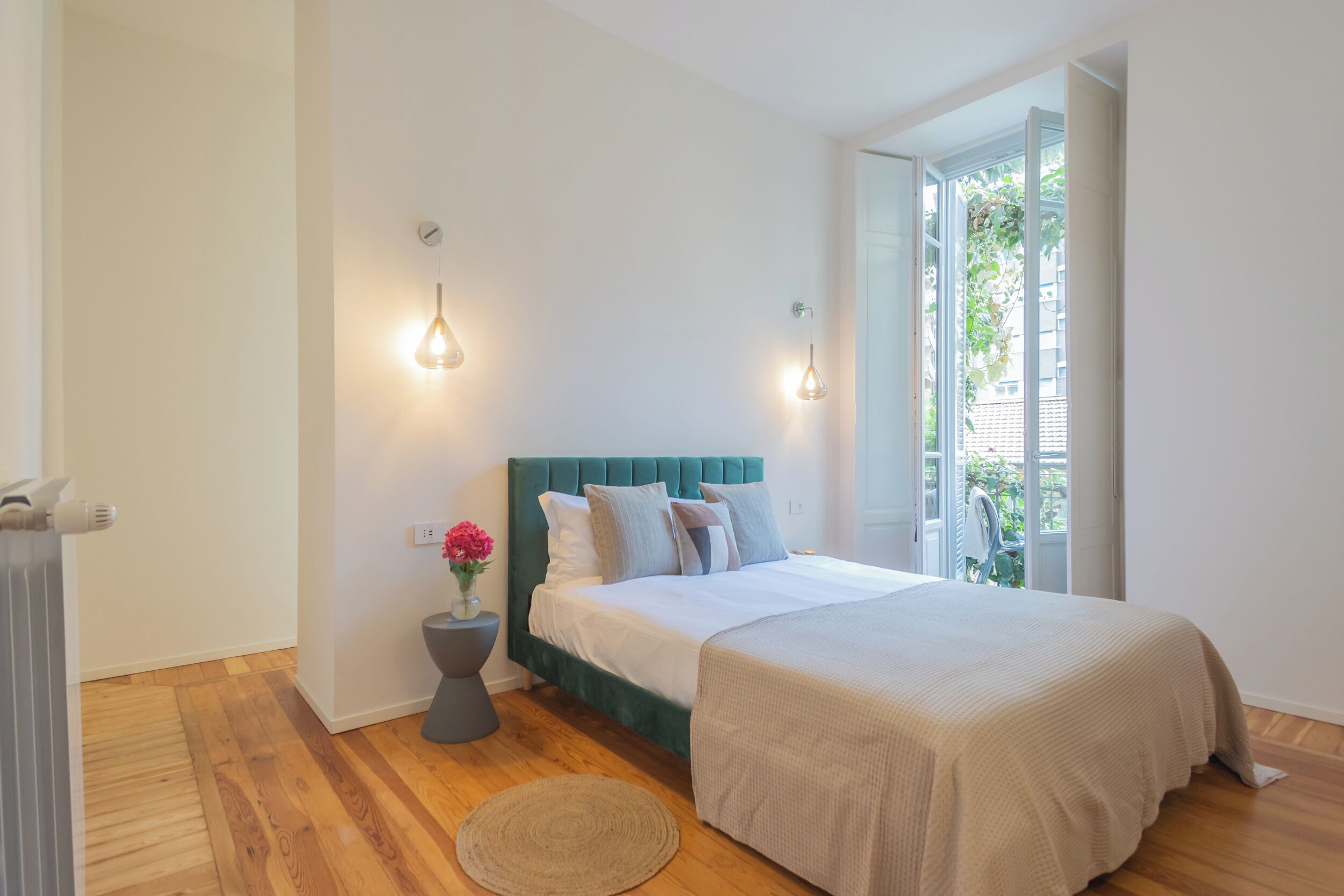 A bedroom with a bed, bedside lamps, a radiator, a round rug, and a window with a view of greenery.