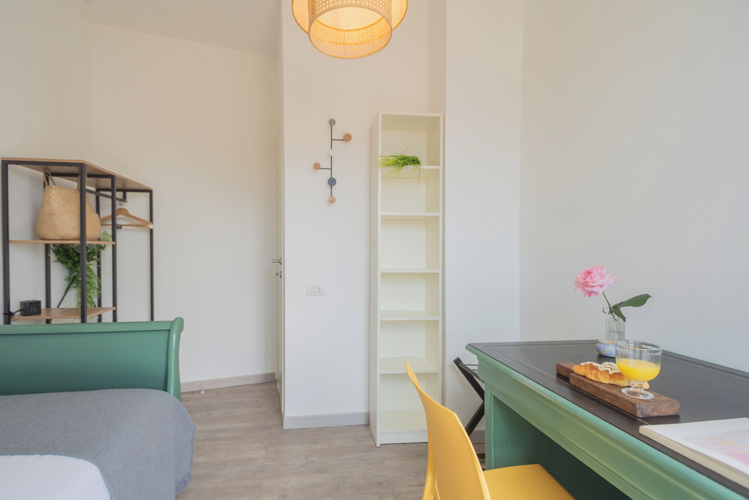 A room with a green bed, a yellow chair, a table with a glass of orange juice, and a white shelving unit.