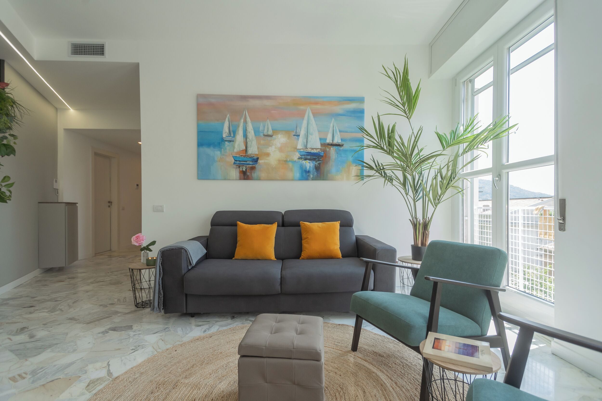 A modern living room with a grey sofa, two yellow cushions, a painting of sailboats, a green armchair, and a large potted plant.