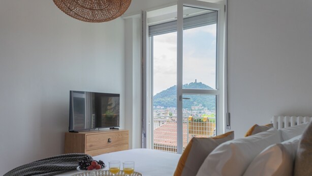 A hotel room with a bed, a TV on a wooden dresser, a tray with fruits and juice, and a large window with a city view. - Como Panorama by Rent all Como (Como)