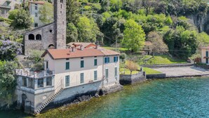 A stone building with a tower, adjacent to a body of water, surrounded by lush greenery and other buildings. - Deriva Apartment on Careno's Beach by Rent all Como (Nesso)