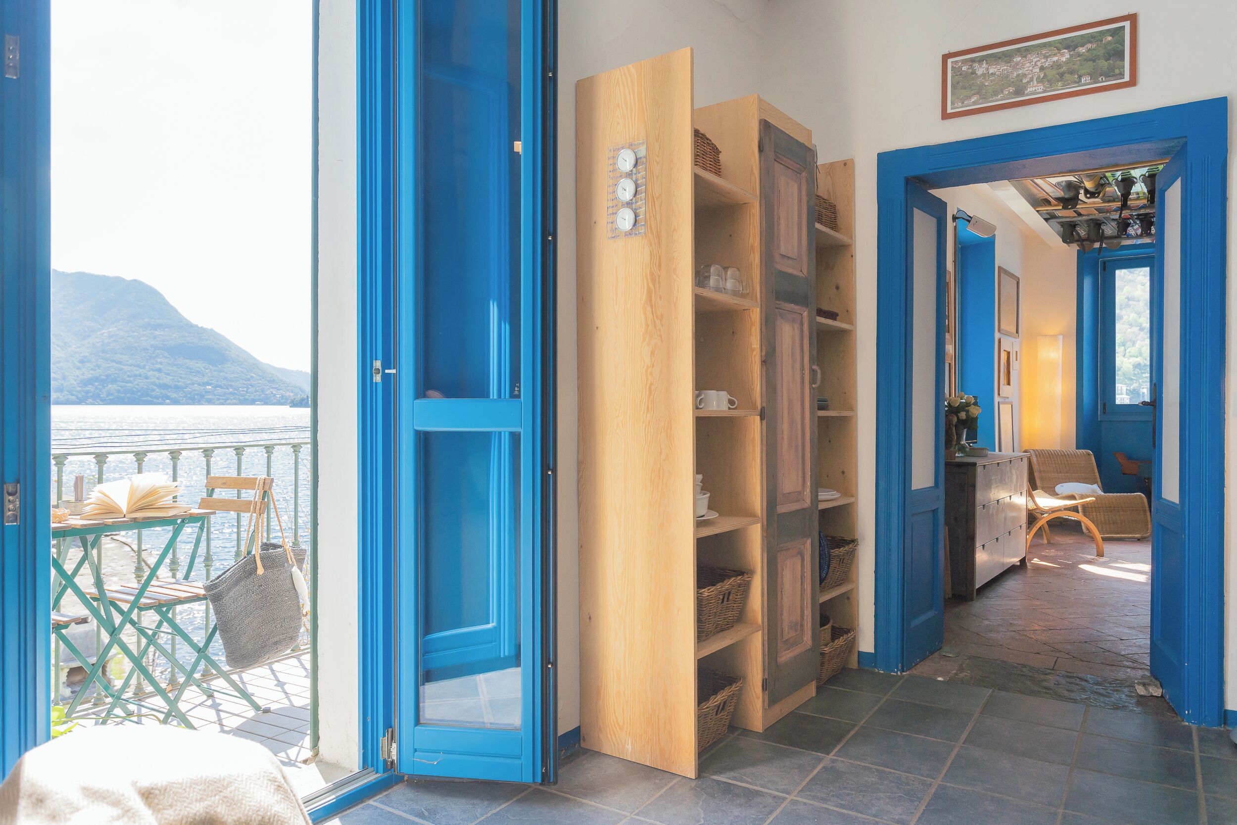Quarto com vista para um corpo de água e montanhas, prateleiras de madeira com cestos, e uma cadeira.