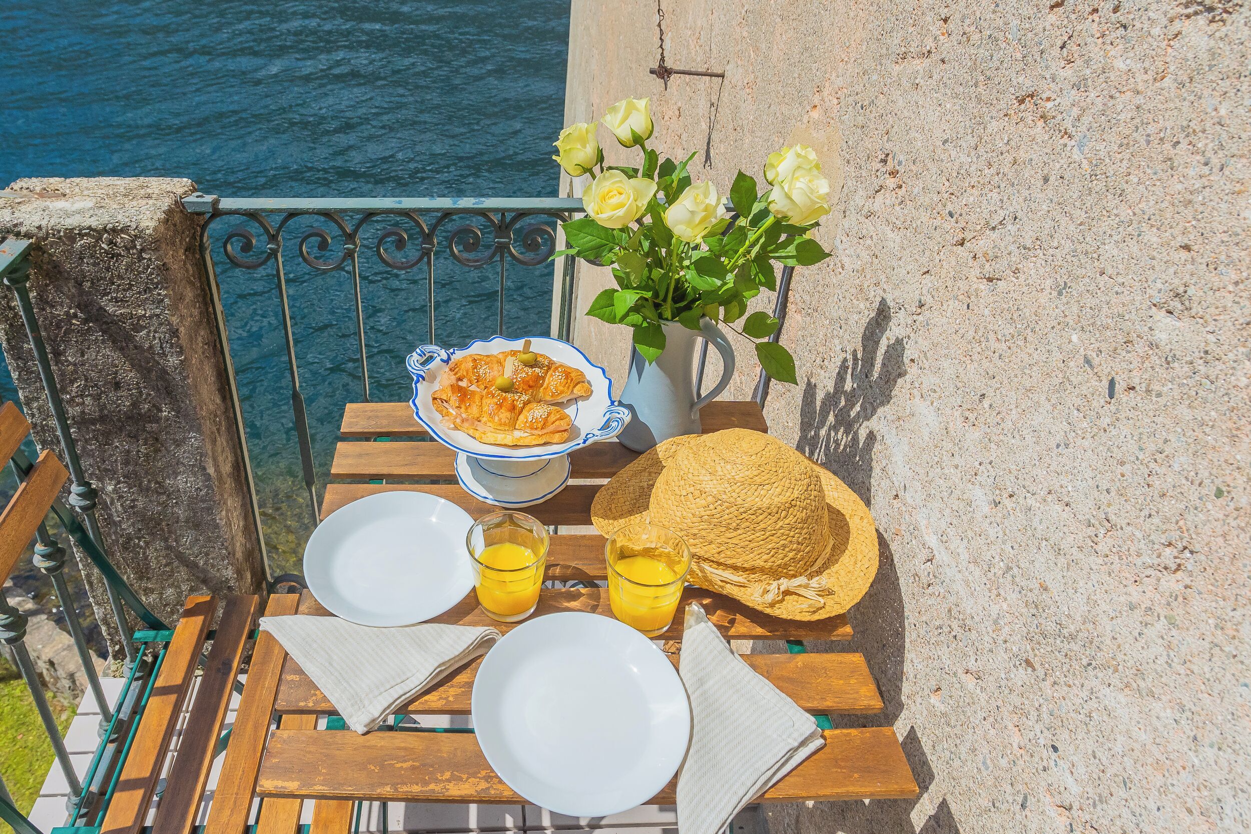 A table set with plates, glasses of orange juice, a basket of bread, and a vase of flowers on a balcony overlooking the sea.