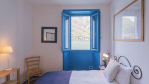 A bedroom with a bed, a chair, a desk, a lamp, and a window with a view of a mountainous landscape. - Deriva Apartment on Careno's Beach by Rent all Como (Nesso)