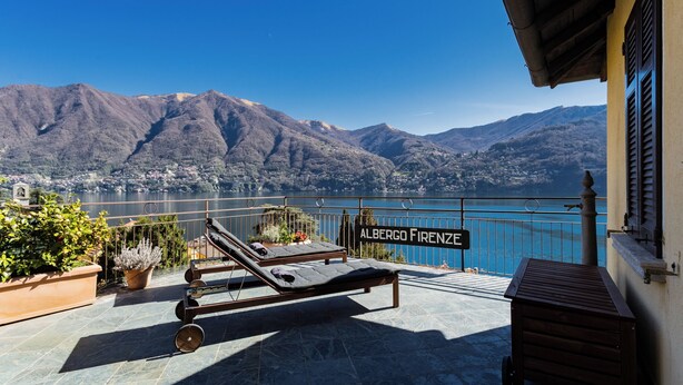 Een terras met twee ligbedden, een bordje met de tekst 'Albergo Firenze' en uitzicht op bergen en een meer.