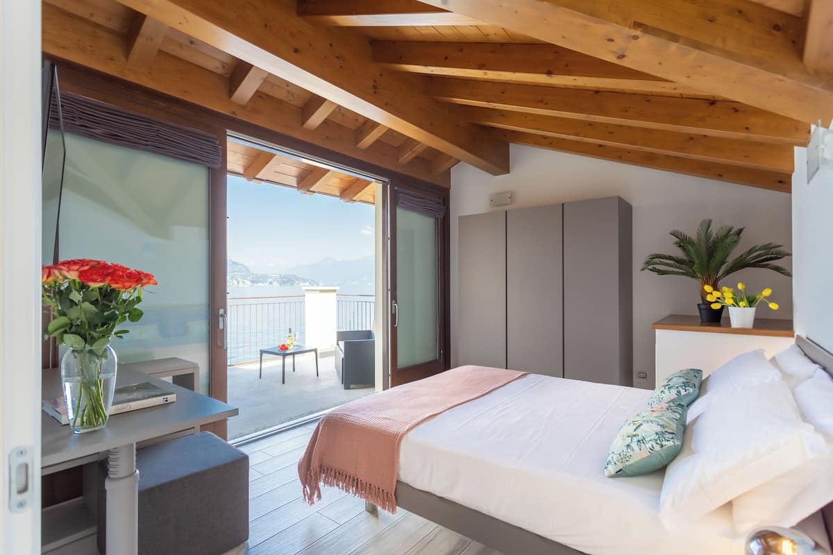 A bedroom with a bed, a bedside table, a chair, a desk, a window, and a balcony with a view of the mountains.