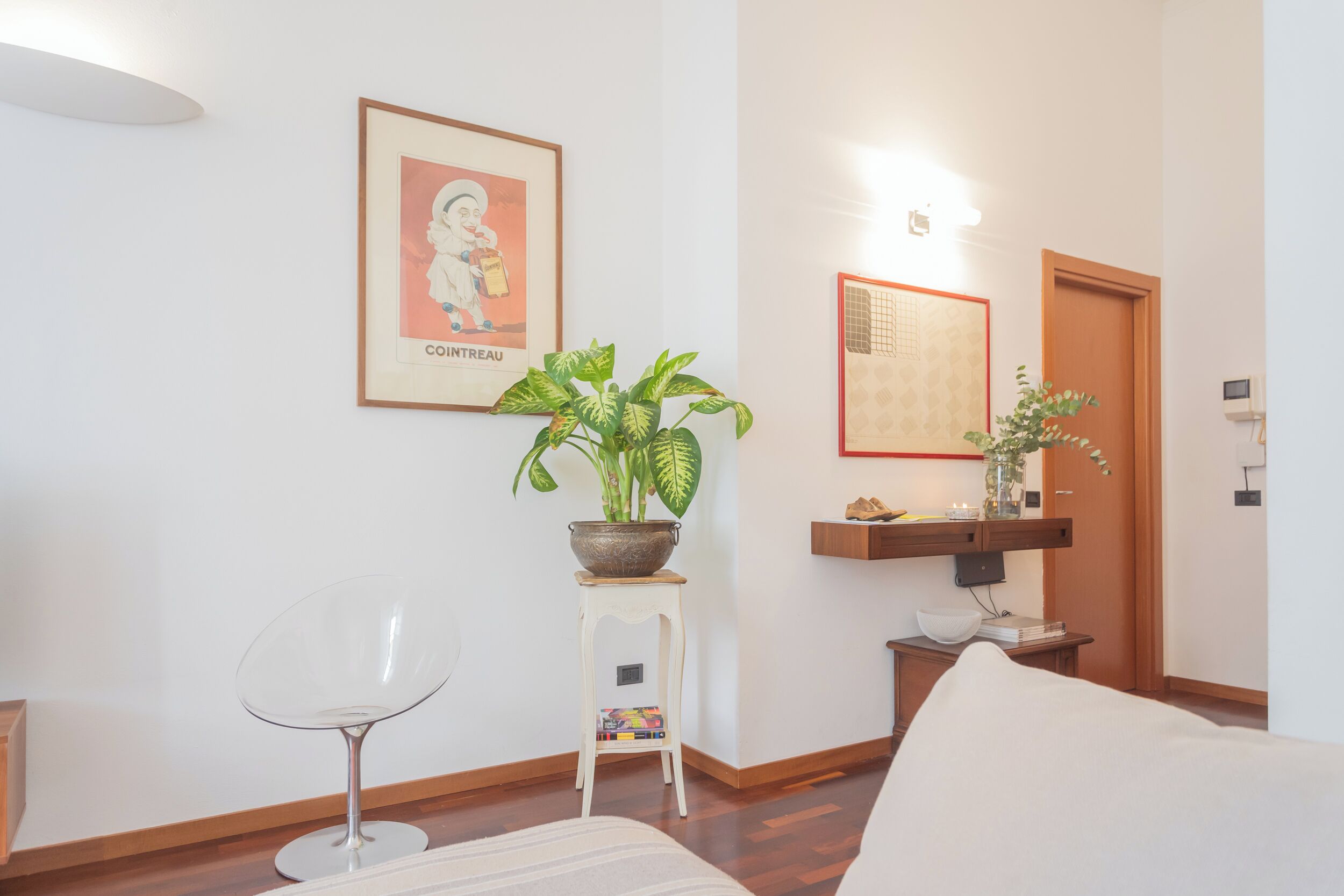 A modern living room with a clear glass chair, a sofa, a potted plant, a framed picture, and a wooden shelf with books and a vase.
