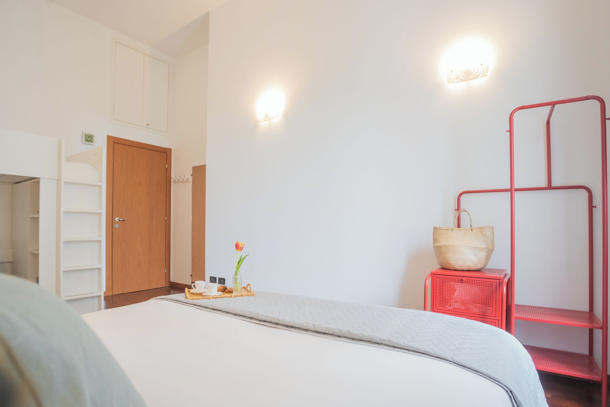 Une chambre d’hôtel avec un lit, une armoire en bois, une étagère en métal rouge et un luminaire fixé au mur.