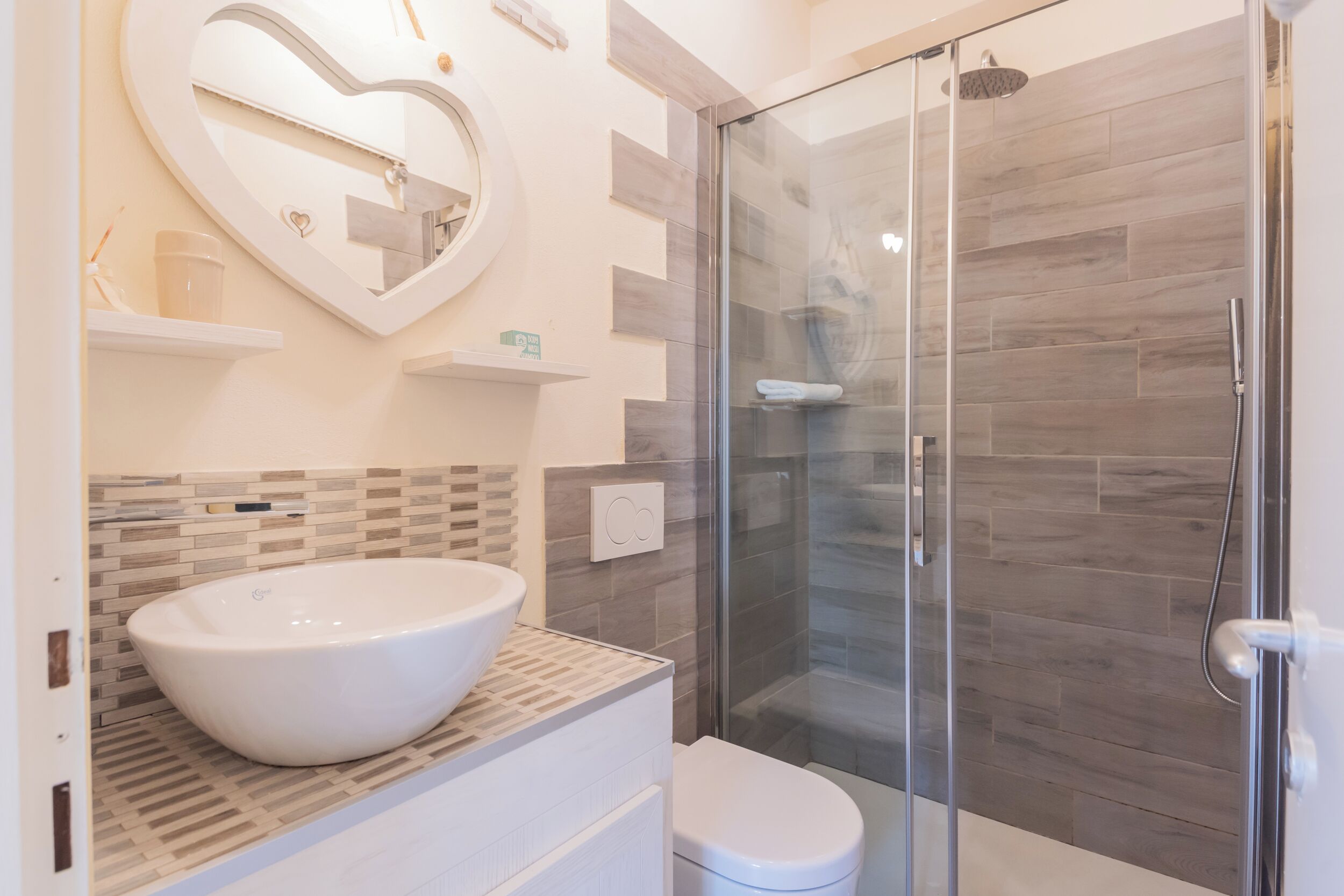 Une salle de bain moderne avec un grand lavabo blanc sur pied, une douche à parois de verre et un miroir en forme de cœur accroché au mur.